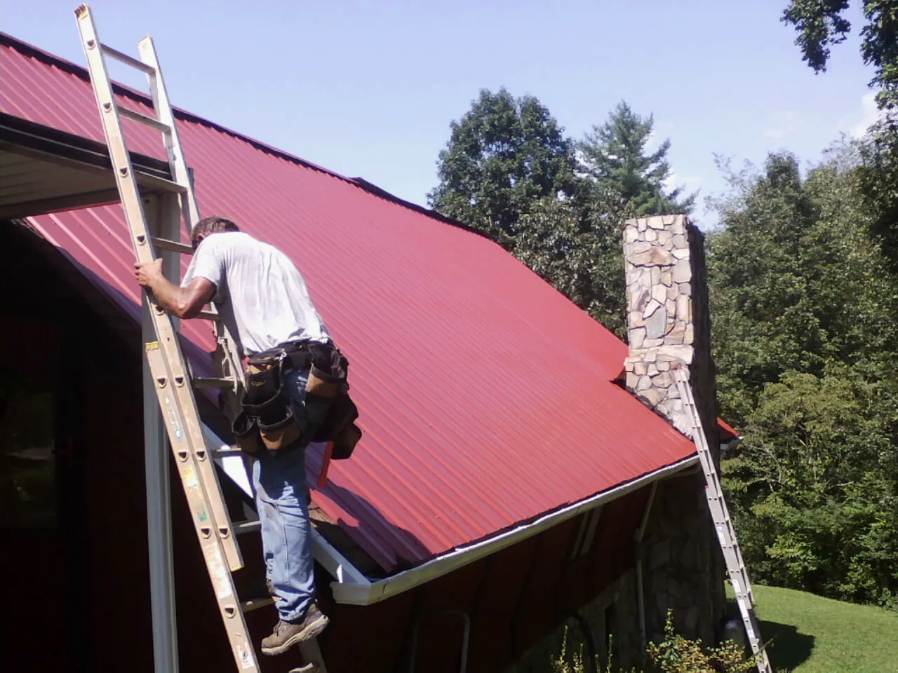 Roofer on a ladder beside a red metal roof and stone chimney — Soffit & Fascia Repair by Roofing Brothers in Alta